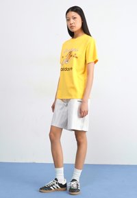 A young woman stands in a yellow graphic t-shirt and striped shorts, wearing black and white sneakers on a blue floor against a white wall.