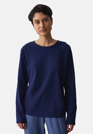 Frau mit kurzen dunklen Haaren, die ein schlichtes marineblaues Langarmshirt und blaue gestreifte Hosen vor einem hellgrauen Hintergrund trägt.