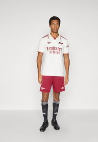 Camisola de futebol branca com detalhes vermelhos e marca "Emirates", combinada com calções vermelhos e meias pretas com riscas e logótipos brancos.