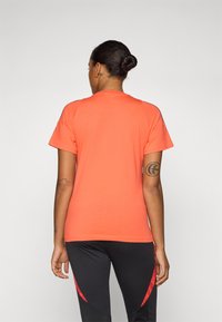 Oranje t-shirt met korte mouwen en een ronde halslijn, gemaakt van een zachte stof. Draagt met zwarte sportlegging met rode accenten.