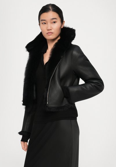 Woman wearing a black leather jacket with a large fur collar and cuffs, paired with a black top and skirt, standing against a plain background.