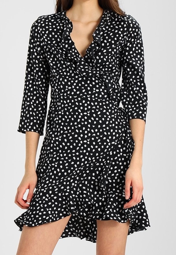 Kobieta w czarnej sukience kopertowej w nieregularne białe groszki, z rękawami do połowy przedramienia, falbanianym dekoltem w serek i asymetrycznym dołem.