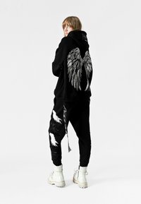 Sudadera negra con diseño de alas metálicas en la parte posterior, combinada con pantalones negros que presentan acentos blancos en forma de alas. El modelo lleva botas blancas con cordones.