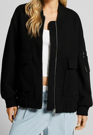 Woman wearing a black zip-up jacket with large front pockets, ribbed cuffs, over a white crop top and light blue pants.