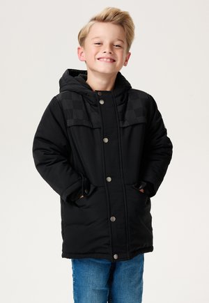 Lachende jongen in een zwarte winterjas met capuchon en een geruit patroon op zijn schouders, met blauwe spijkerbroek, handen in de zakken, staand op een witte achtergrond.