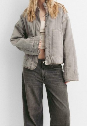 Kvinde iført beige kort quiltet jakke over hvid kort top med sorte vidde jeans mod ens hvid baggrund.