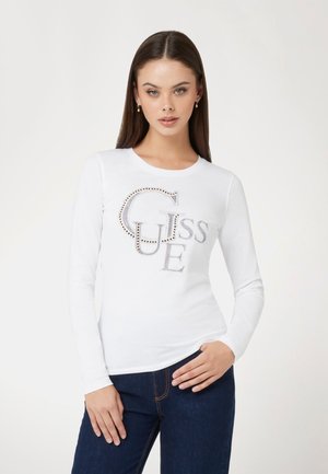 Baltas ilgomis rankovėmis marškinėliai su dideliu logotipu "GUESS", papuoštu kristalais. Minkšta medvilnės medžiaga su prigludusia siluetu.