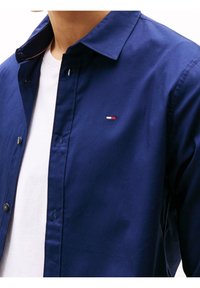 Camisa de algodón azul marino con cuello abotonado, costuras visibles y un pequeño logo con detalles en rojo, blanco y azul en el pecho.