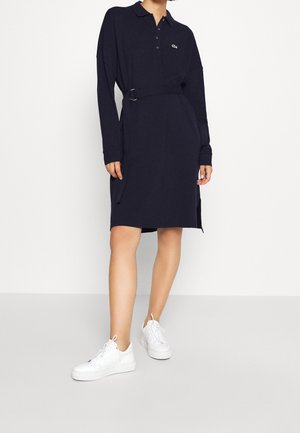 Vrouw draagt een marineblauwe polo jurk met lange mouwen en ceintuur, gecombineerd met witte sneakers, staand op een lichtgrijze vloer tegen een witte achtergrond.