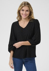 Mustmust tekstuuriga must blouse, madala V-kaelusega, balloonikujuliste varrukatega ja auguliste mustritega. Kombineeritud siniste teksadega, et luua juhuslik välimus.