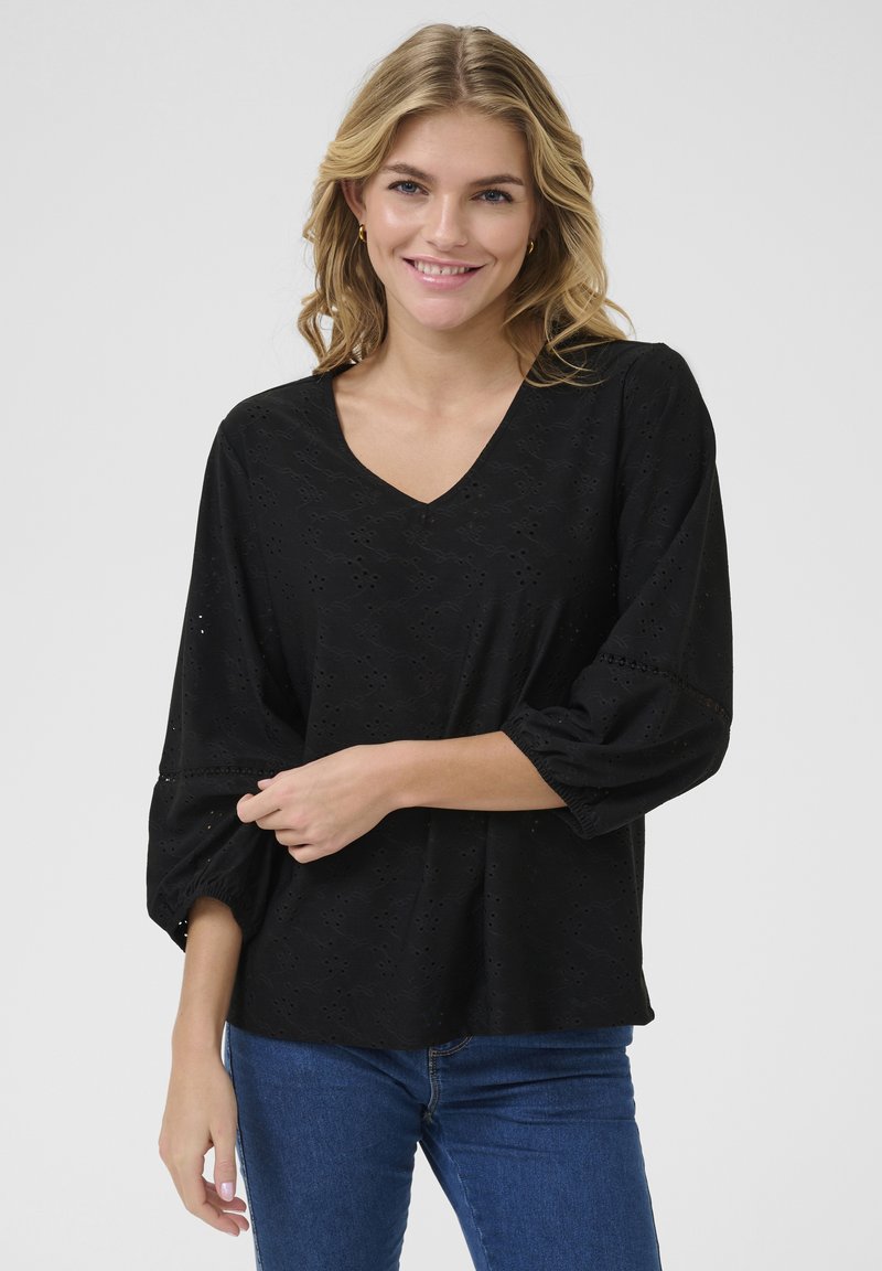 Zwarte blouse met textuur, een V-hals, ballonmouwen en opengewerkte patronen. Gecombineerd met een blauwe jeans voor een casual uitstraling.