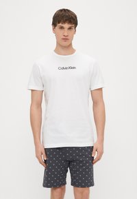 Bela bombažna majica s črnim logotipom "Calvin Klein", kombinirana s temno sivimi kratkimi hlačami z belim vzorčnim dizajnom.