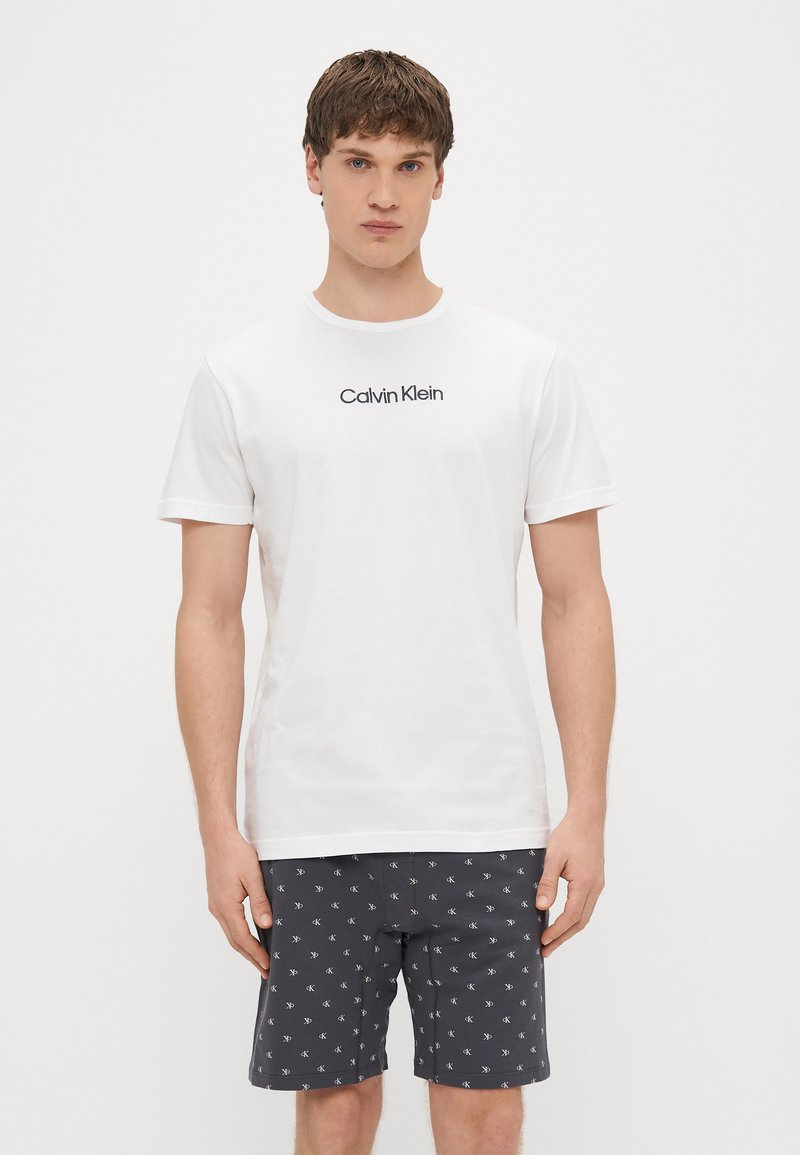 Bela bombažna majica s črnim logotipom "Calvin Klein", kombinirana s temno sivimi kratkimi hlačami z belim vzorčnim dizajnom.