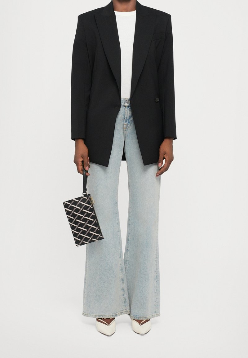 Personne portant un blazer noir, un haut blanc, un jean large bleu clair, des chaussures blanches pointues et tenant un sac bracelet noir à motifs.