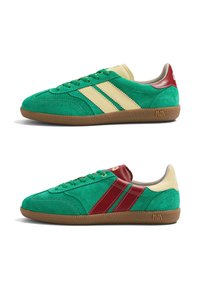 Deux baskets en daim vert présentées de côtés opposés, avec des bandes beige et rouge, des semelles en caoutchouc marron, et un design bas.