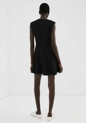 Žena stojaca chrbtom, oblečená v čiernych šatách bez rukávov a bielych tenisových topánkach na jednoduchom bielom pozadí.