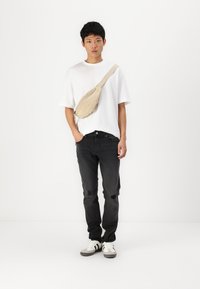 Beige høydeveske brukt over en hvit T-skjorte, kombinert med svarte jeans med slitt detaljer og hvite sneakers med grå aksenter.