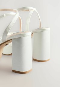 Bílé sandály na podpatku s hladkým saténovým povrchem, jež mají blokový podpatek a design s křížovými pásky. Doplňky zahrnují béžovou podrážku.