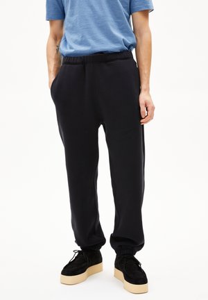 Persoon draagt zwarte joggers met elastische taille en cuffed enkels, zwarte platform schoenen en een blauw gestreept shirt met korte mouwen.