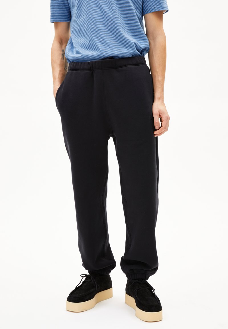 Personne portant un pantalon de jogging noir à taille élastique avec des chevilles resserrées, des chaussures à plateforme noires et une chemise à manches courtes rayée bleue.