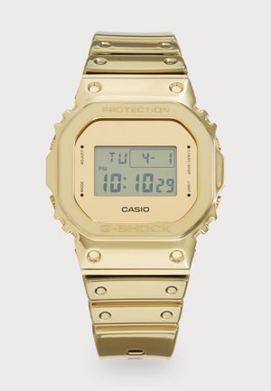 Auksinis skaitmeninis laikrodis su kvadratiniu cypiu, turintis pilką LCD ekraną. Lygaus metalo apyrankė su studais, pažymėtais mygtukais ir iškilmingu prekės ženklu.