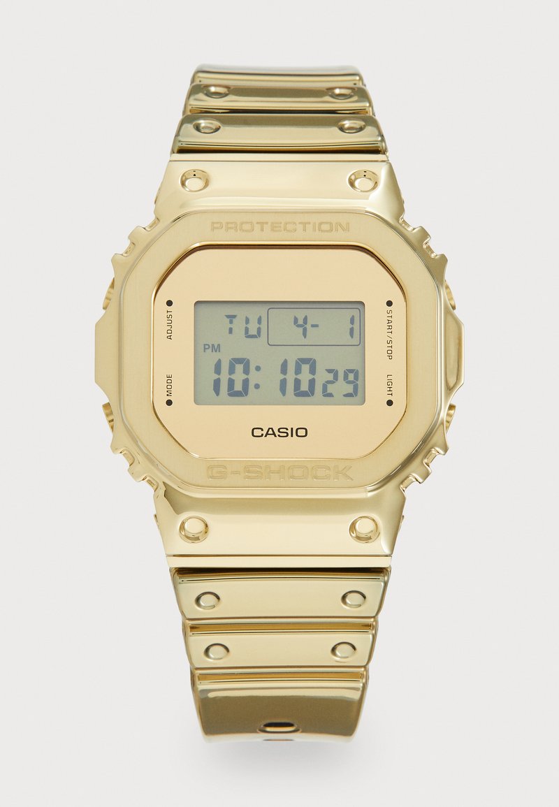 Orologio digitale dorato con quadrante quadrato, dotato di un display LCD grigio. Bracciale in metallo liscio con borchie, pulsanti etichettati e nome del marchio in rilievo.