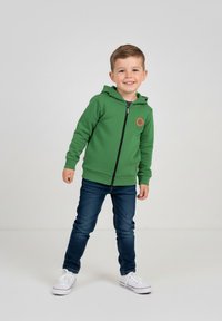 Zielona bluza z kapturem na zamek błyskawiczny, wykonana z teksturowanej tkaniny, z okrągłą naszywką z logo, zestawiona z ciemnoniebieskimi dżinsami i białymi trampkami.