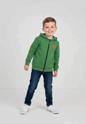 Zielona bluza z kapturem na zamek błyskawiczny, wykonana z teksturowanej tkaniny, z okrągłą naszywką z logo, zestawiona z ciemnoniebieskimi dżinsami i białymi trampkami.