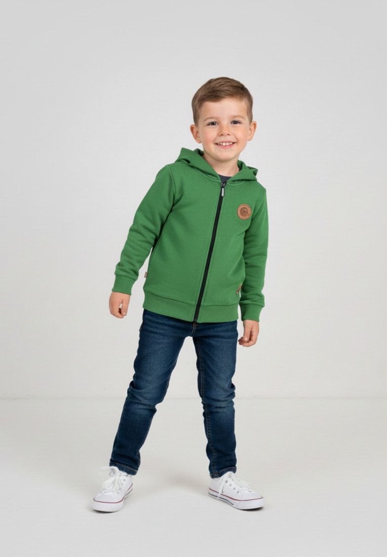 Zielona bluza z kapturem na zamek błyskawiczny, wykonana z teksturowanej tkaniny, z okrągłą naszywką z logo, zestawiona z ciemnoniebieskimi dżinsami i białymi trampkami.