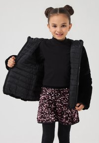 Giacca a vento nera con motivo a quadri, indossata sopra un top nero e una gonna rosa lucida e con paillettes. La giacca ha una chiusura con zip e una consistenza morbida.