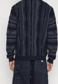 Svetr s texturovaným vzorem námořnických modrých a šedých pruhů, žebrovanými manžetami a lemem, vyrobený z měkkého pleteného materiálu.