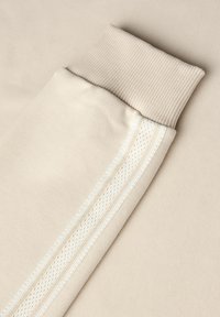 Tessuto di felpa beige con polsini a coste. Presenta una striscia verticale bianca con un motivo a rete testurizzata lungo il lato.