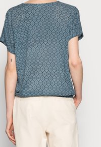 Top de manga corta azul con un diseño geométrico, fabricado en una tela suave y ligera, y con un corte holgado. Combinado con pantalones beige.