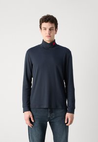 Maglietta a maniche lunghe blu navy con collo alto e dettaglio in etichetta rossa, realizzata in un tessuto liscio ed elastico, abbinata a jeans in denim scuro.