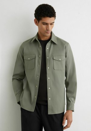 Mees, kes kannab oliivirohelise värvi nööpidega särki musta topi peal, seisab valge tekstuurse seina taustal, vaatab alla ja hoiab käed lõdvestunult.