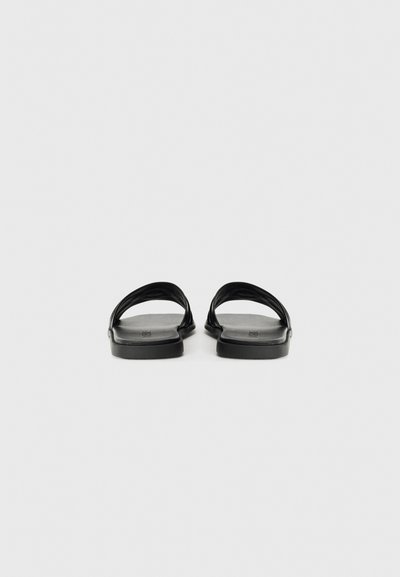 Svarta platta sandaler med en minimalistisk design, med två böjda, överlappande remmar. Sulan är slät med en matt yta.