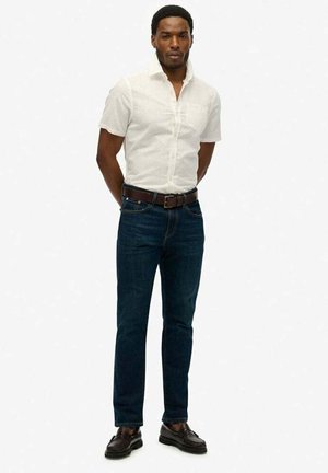 Hvid kortærmet skjorte med knap, mørkeblå slim-fit jeans, brun læderbælte og mørke sko. Ensfarvet baggrund.