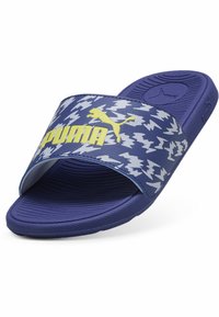 Blå slide-sandal med en texturerad sula, med en djärv gul "PUMA"-logotyp på ett mönstrat rem med ett marinblått och vitt zigzag-mönster.