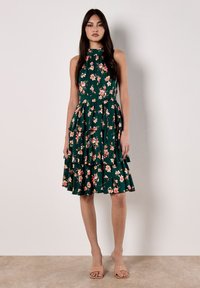 Robe florale verte avec un col montant, des volants superposés et des motifs de fleurs roses. Portée avec des sandales nude. Texture de tissu lisse.