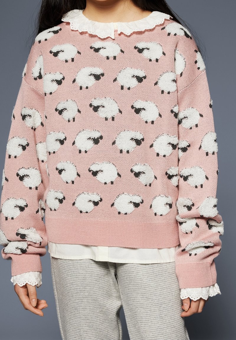 Enfant portant un pull rose avec un motif de mouton blanc, un col et des poignets volantés blancs, et un pantalon à carreaux gris sur fond gris.
