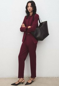 Žena stojící se zkříženýma rukama, oblečená v burgundském obleku, s černými slingback podpatky a nesoucí velkou černou koženou kabelku na rameni.