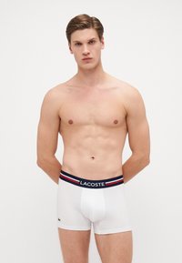 Weiße Baumwoll-Boxershorts mit einem marineblauen Bund, der rot-weiße Streifen aufweist. Lacoste-Logo auf der linken Seite, glatte Textur, taillierte Passform.