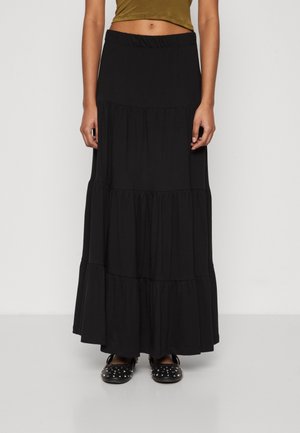 Femme portant une longue jupe noire à volants, un crop top vert olive et des ballerines noires avec des clous argentés, debout devant un fond uni.