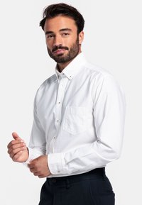 Man met donker haar en baard, gekleed in een wit knoopshirt en donkere broek, die zijn manchet aanpast tegen een plain witte achtergrond.