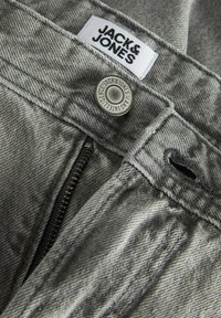 Hallatav sissevõte hallidest teksapükstest nähtava luku, metallist nupuga, millele on graveeritud „Jack & Jones“, ja valge kangast brändisildiga.