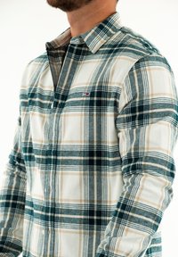 Camisa de franela a cuadros en blanco, verde y marrón. Cuenta con cuello abotonado, mangas largas y un pequeño logo en el pecho. Textura suave.