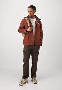 Pruun veekindel jakk musta kraega, hall särk, pruunid püksid ja khaki matkasussid. Kujutis kannab beežikat seljakotti.