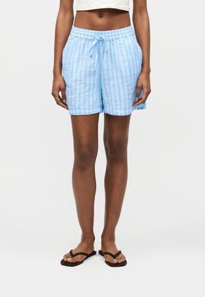 Γυναίκα που φοράει ανοιχτό μπλε ριγέ σορτς με κορδόνι, καφέ σαγιονάρες και λευκό κοντό τοπ, στέκεται μπροστά σε απλό φόντο.