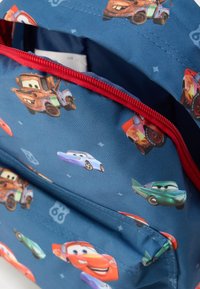 Σακίδιο με μπλε ύφασμα που απεικονίζει πολύχρωμα καρτούν αυτοκίνητα. Το κόκκινο φερμουάρ ξεχωρίζει από το μπλε φόντο, το οποίο περιλαμβάνει μοτίβα οχημάτων και διακοσμητικά στοιχεία.