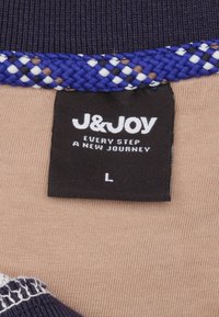 Etiket met 'J&JOY' logo, zwarte achtergrond; beige stof met gebreide marineblauwe en witte rand; maat 'L'; bevat 'IEDERE STAP EEN NIEUWE REIS.'